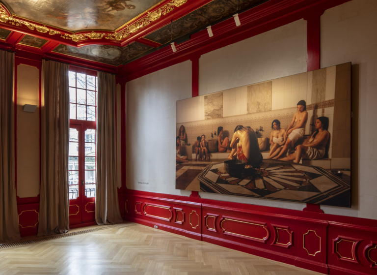 Ornate historic room with deep red walls, gold detailing, and a painted ceiling, featuring tall windows, a fireplace with mirror, and contemporary artworks hanging on the walls.